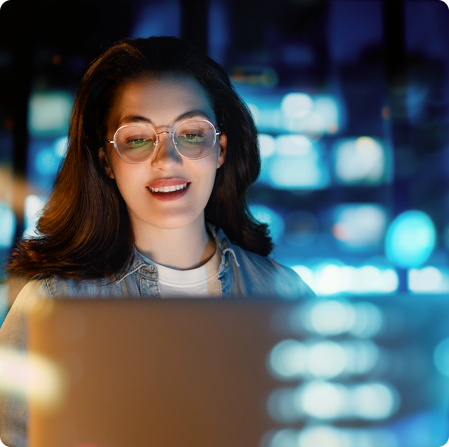 Woman with glasses smiling while using laptop at night with city lights in background.