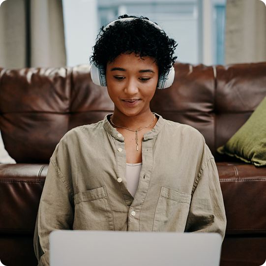 Person wearing headphones and beige shirt using a laptop on a couch.